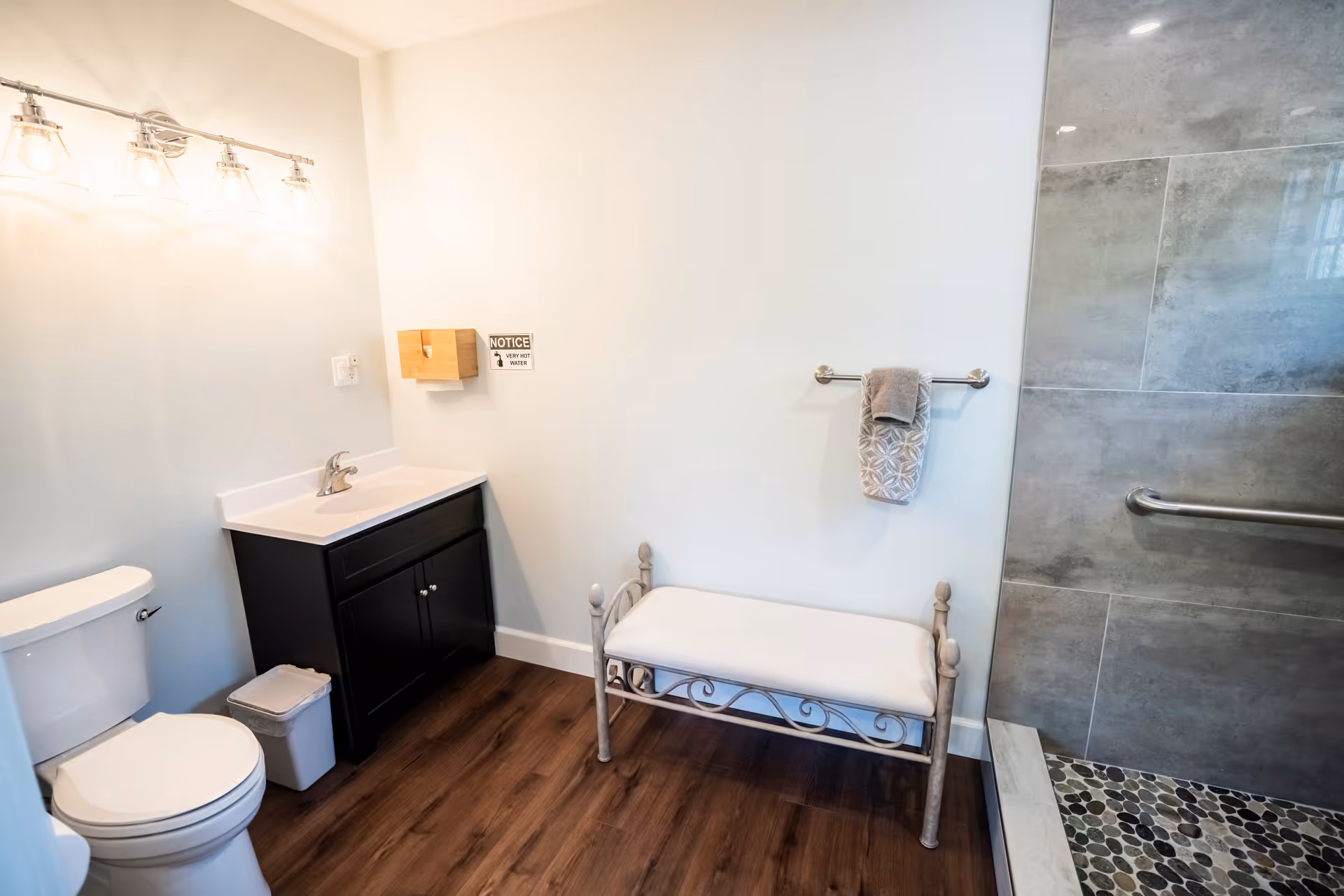A clean and modern bathroom featuring a white toilet, a black vanity with a white sink, a wall-mounted light fixture with four bulbs, a small trash bin, a cushioned bench with decorative metal legs, a towel rack with a folded towel, and a walk-in shower with gray tiles and pebble flooring.