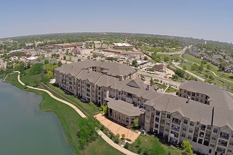 Aerial view of a large senior living facility building situated next to a lake with a walking path along the water. The building has multiple floors with balconies and is surrounded by green lawns and trees. In the background, there is a suburban area with roads, houses, and commercial buildings under a clear sky.