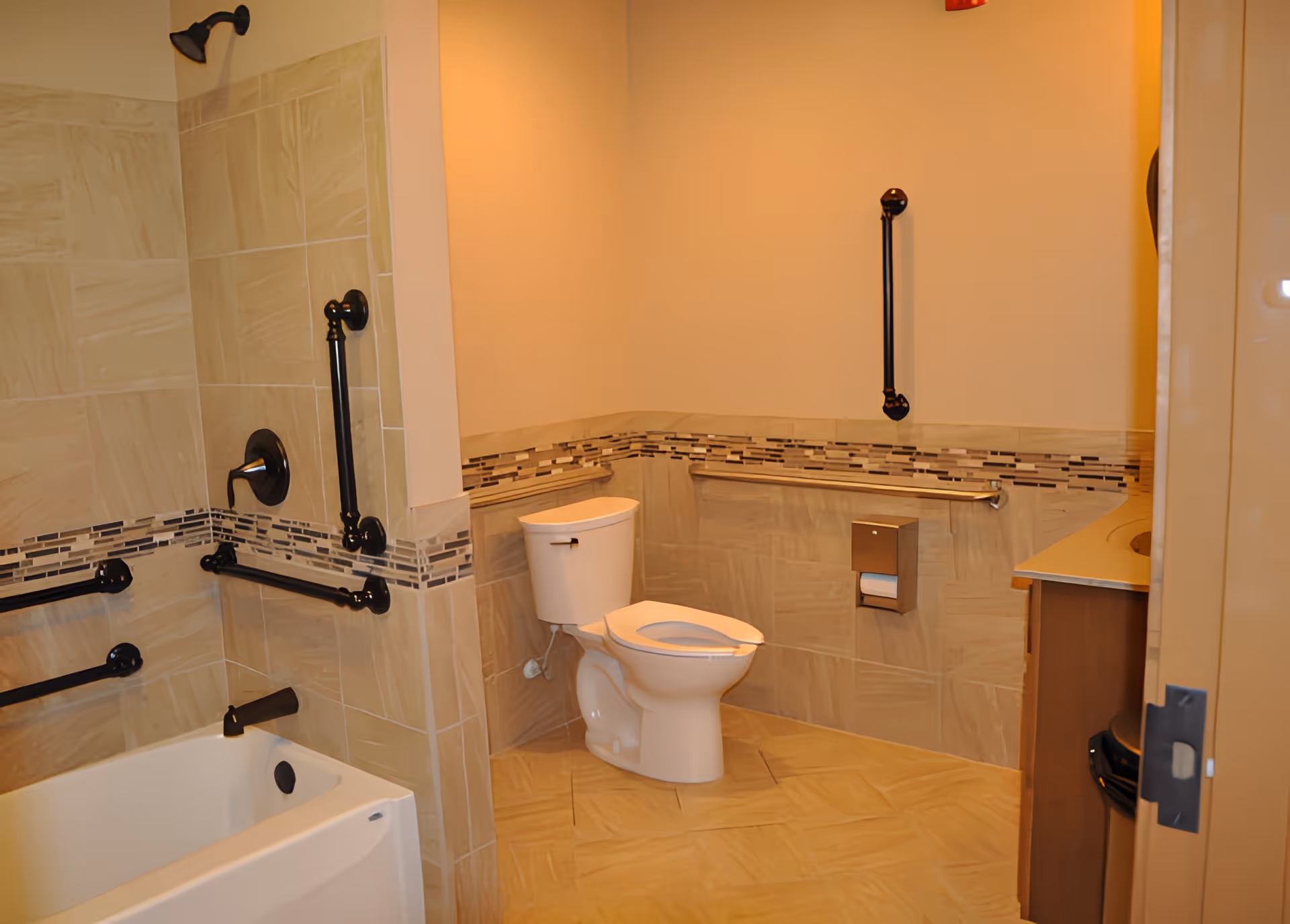 A bathroom with beige tiled walls and floor, featuring a white toilet with an open lid, a bathtub with black fixtures and grab bars, and a sink countertop partially visible on the right side.