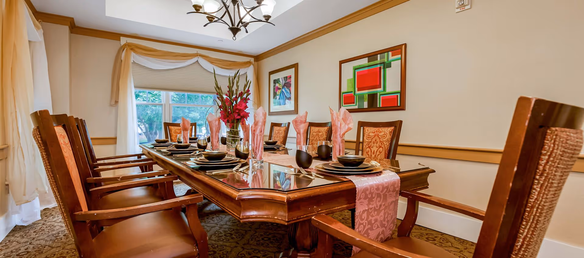 Elegant dining room with a long wooden table set for a meal, surrounded by upholstered chairs, floral centerpiece, and framed artwork.