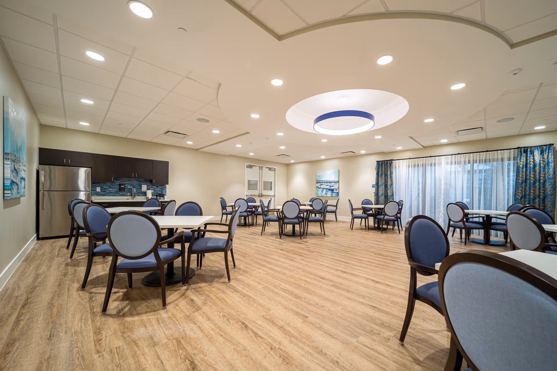 A spacious dining room with multiple round tables and blue cushioned chairs. The room features wood flooring, a modern ceiling light fixture, and a kitchenette area with dark cabinets and a stainless steel refrigerator. Large windows with blue patterned curtains allow natural light to enter the room.