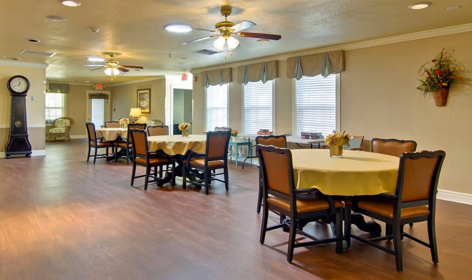 A spacious dining room with round tables covered with yellow tablecloths, each surrounded by four brown cushioned chairs. The room has large windows with blinds and valances, a wooden floor, ceiling fans with lights, and a tall grandfather clock near the entrance. There are also some floral decorations on the walls and tables.
