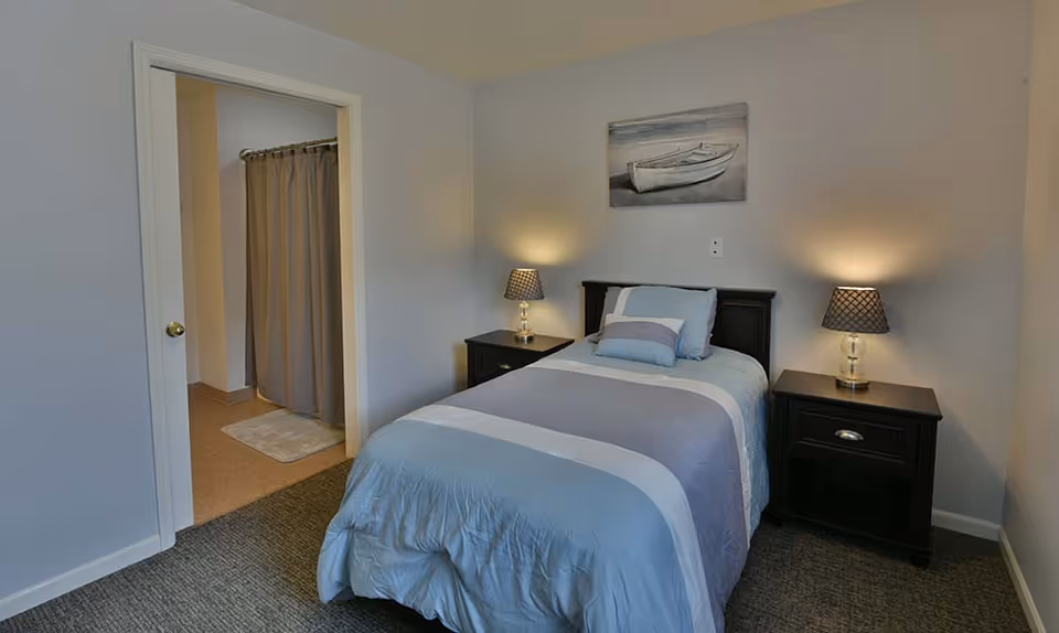 A small bedroom with a single bed made with blue and gray bedding, flanked by two dark wooden nightstands each with a lamp. A painting of a boat hangs on the wall above the bed. An open doorway reveals a bathroom with a shower curtain and bath mat.