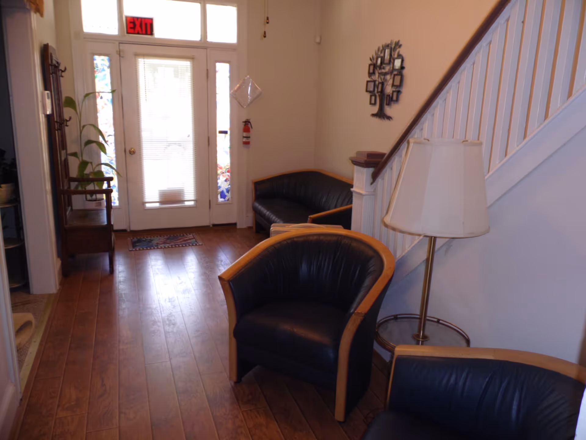 A small waiting or sitting area with wooden flooring, two black leather chairs with wooden frames, a black leather loveseat, a floor lamp with a white shade, a wooden coat rack, a plant, and a white door with glass panels and an illuminated red exit sign above it.