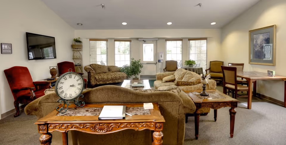 A cozy living room area in a senior living facility with multiple upholstered sofas and armchairs arranged around wooden tables. A large clock sits on one of the tables, and a flat-screen TV is mounted on the wall. There are large windows and a door letting in natural light, with plants placed near the windows. A restroom sign is visible on the wall.