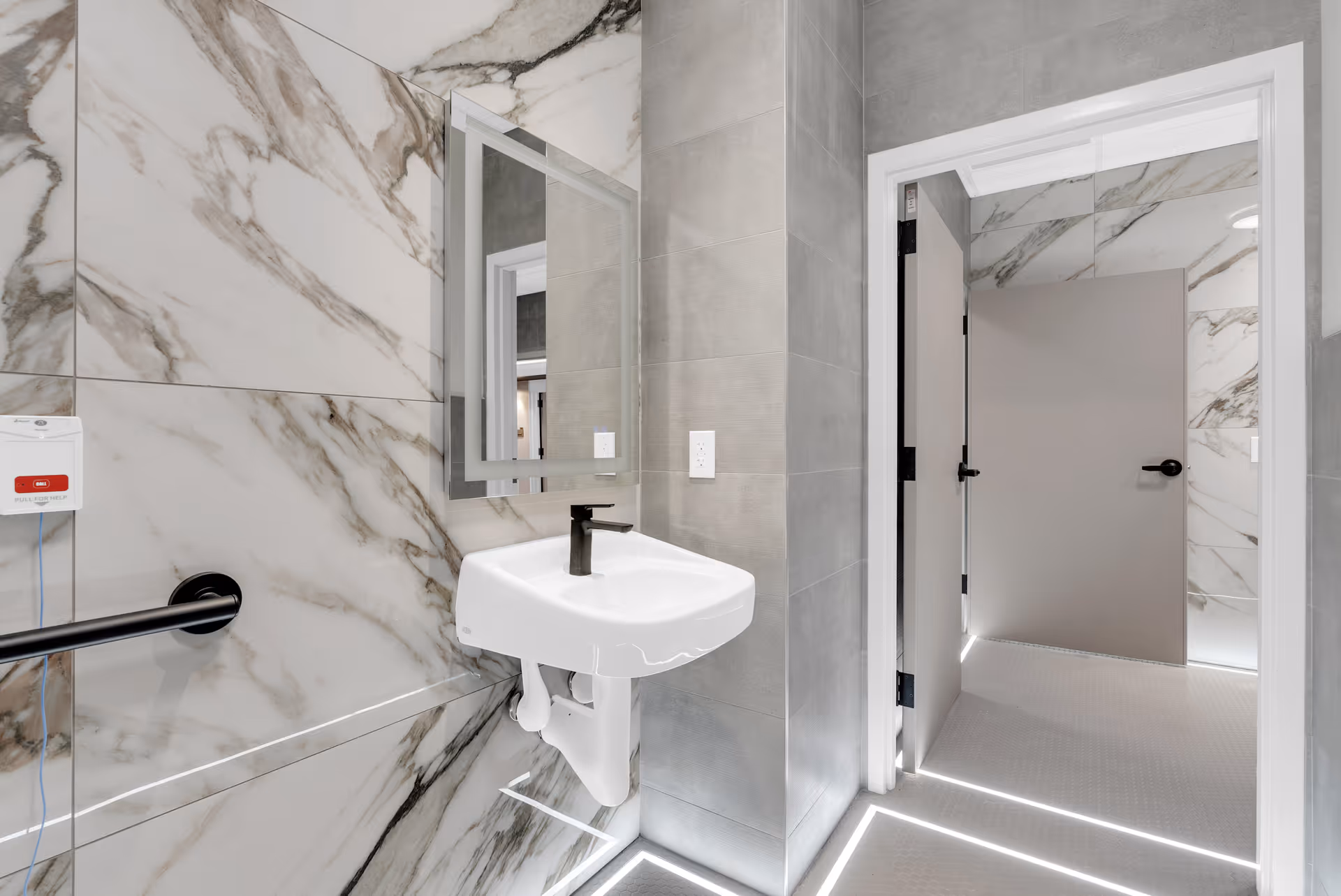 Modern accessible bathroom with marble-patterned walls, wall-mounted sink and mirror, a grab bar, and an open doorway leading to a hallway.