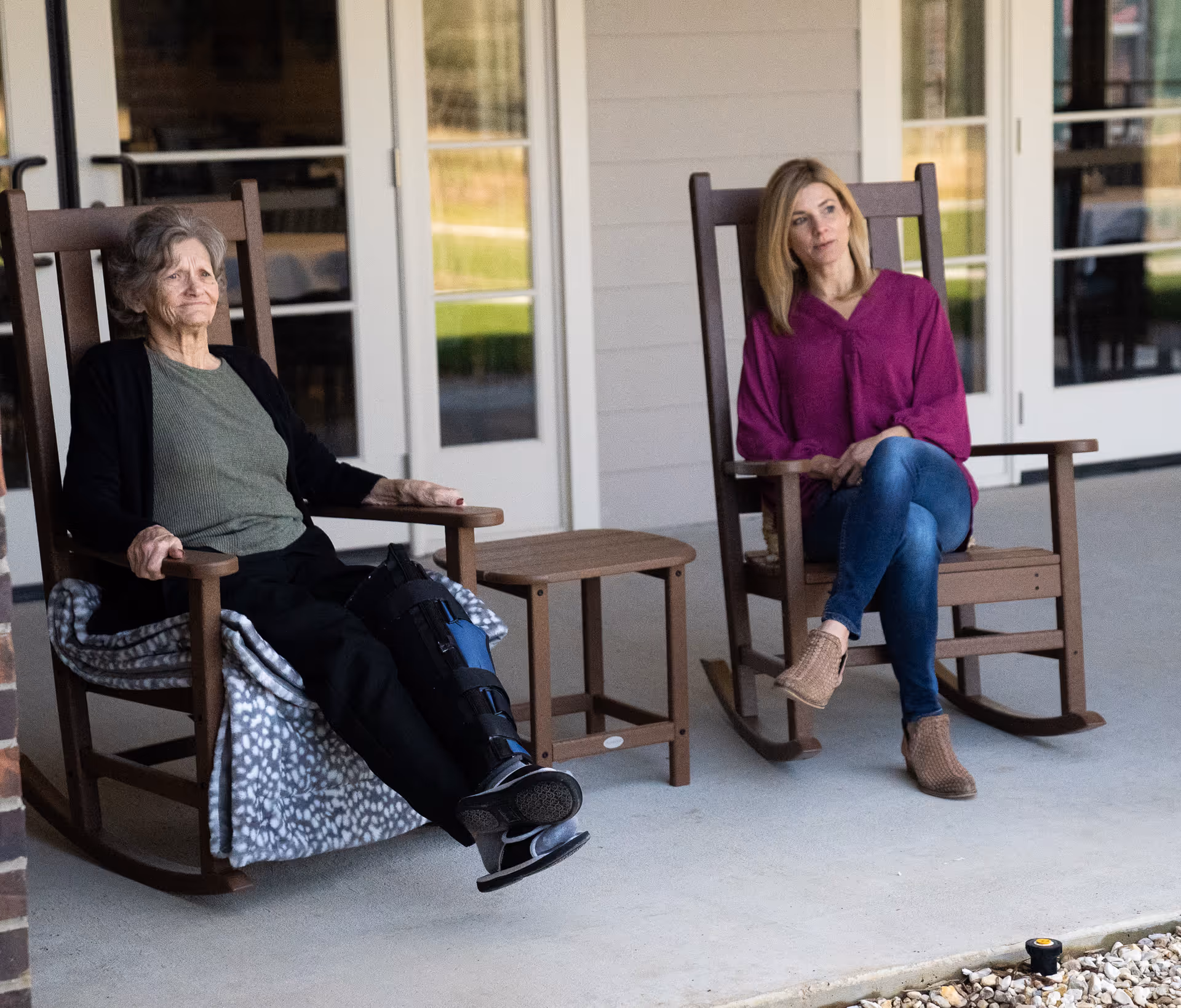 An elderly woman wearing a leg boot and a younger woman sit on wooden rocking chairs on a covered porch outside a facility.