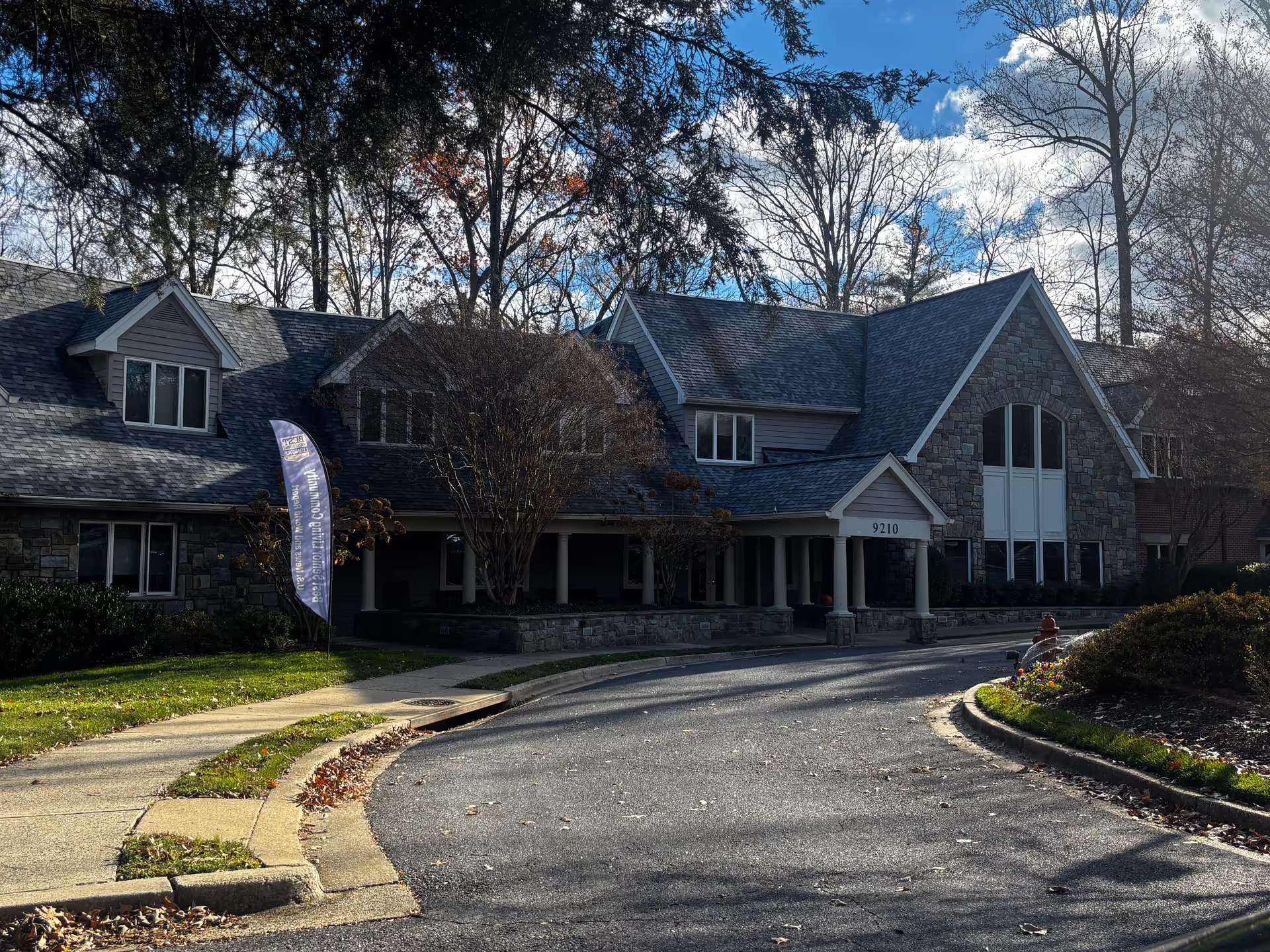 Exterior view of Byron House, a senior living facility, showing a large stone and siding building with a peaked roof, multiple windows, and a covered entrance with columns. The driveway curves in front of the entrance, and there are trees and bushes around the building under a partly cloudy sky.