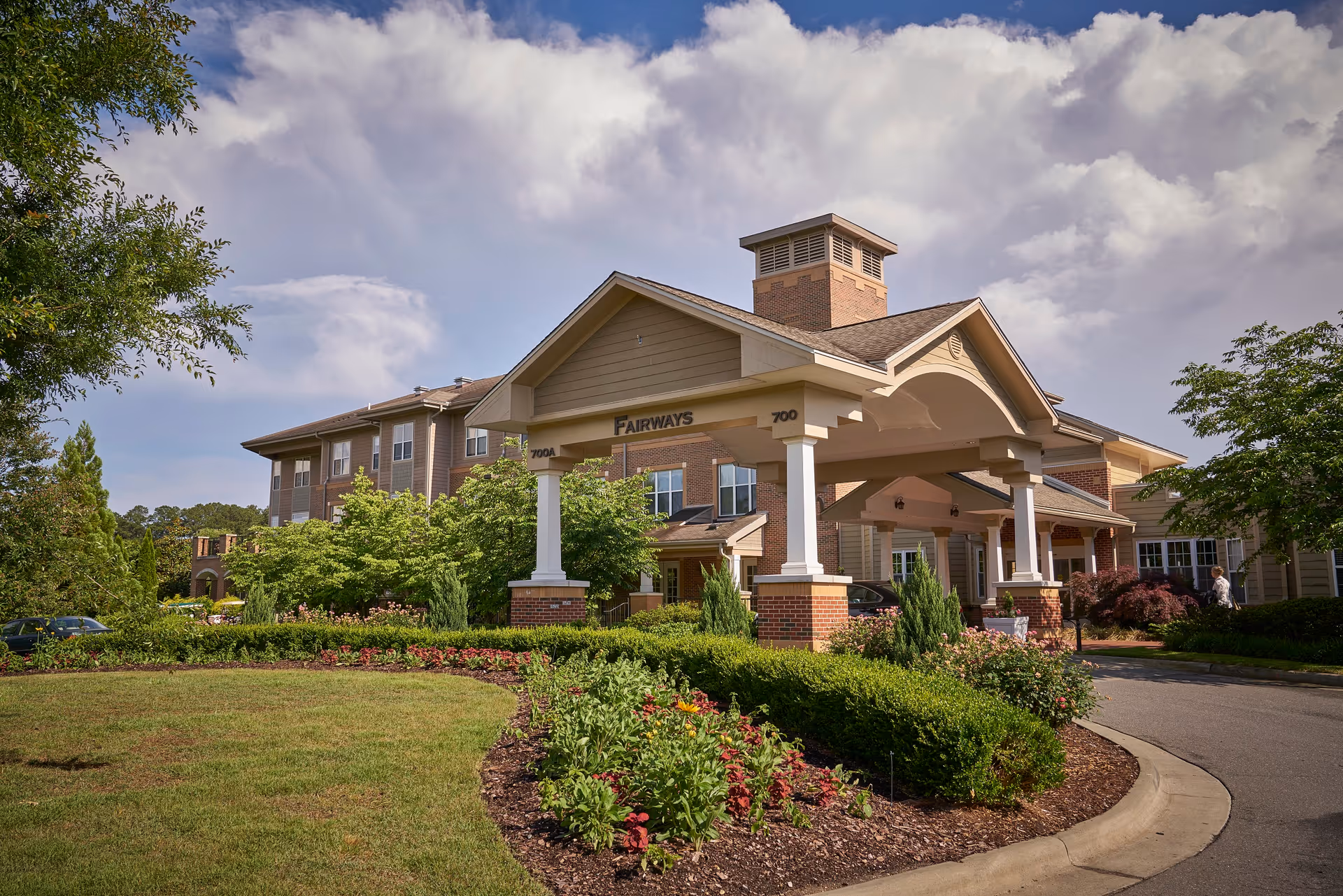 Exterior view of a senior living facility named Carolina Meadows showing a covered entrance labeled 'Fairways' with well-maintained landscaping including bushes, flowers, and trees under a partly cloudy sky.