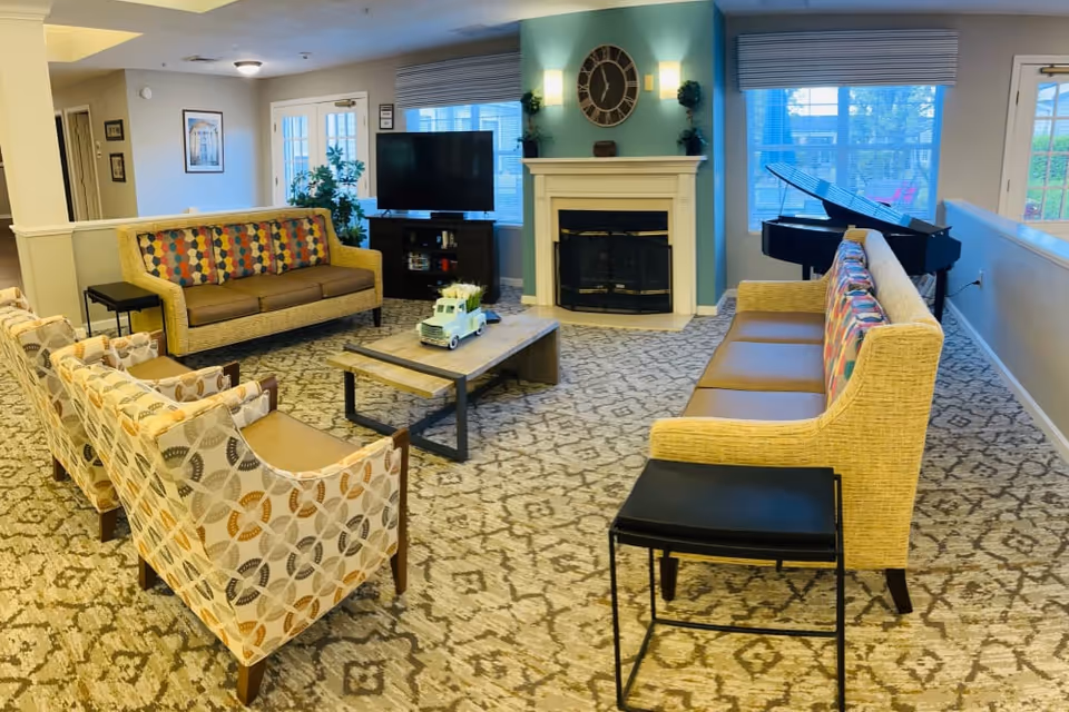 A cozy living room area with patterned carpet, two sofas with colorful cushions, three patterned armchairs, a wooden coffee table with a decorative truck, a fireplace with a large clock above it, a flat-screen TV on a stand, and a grand piano near large windows with blinds.