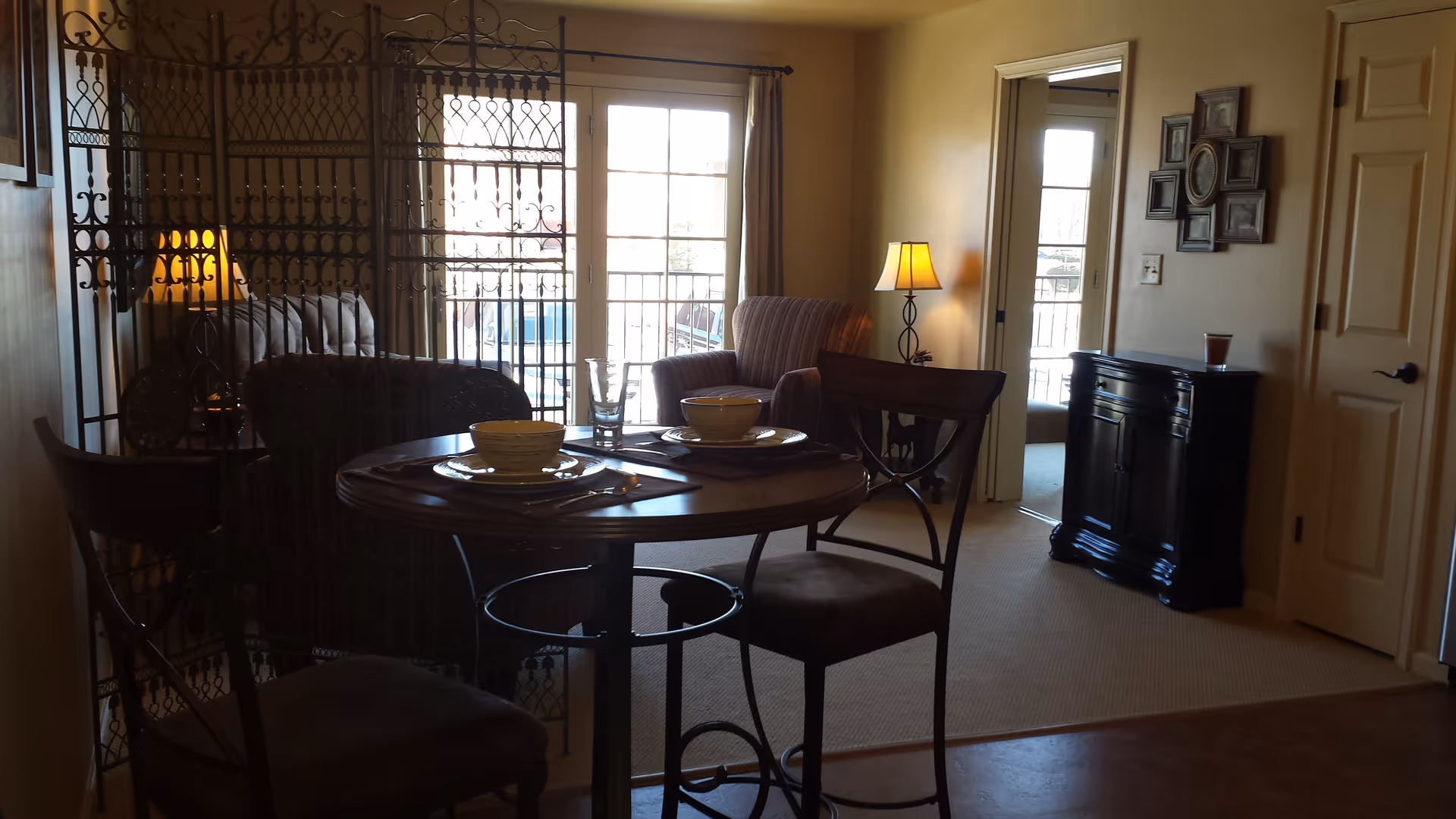Interior view of a senior living facility showing a small round dining table set for two with bowls and plates, two chairs, a decorative metal room divider, an armchair with a floor lamp beside it, a black cabinet with a glass on top, and a door leading to another room. Large windows with curtains allow natural light to enter the space.
