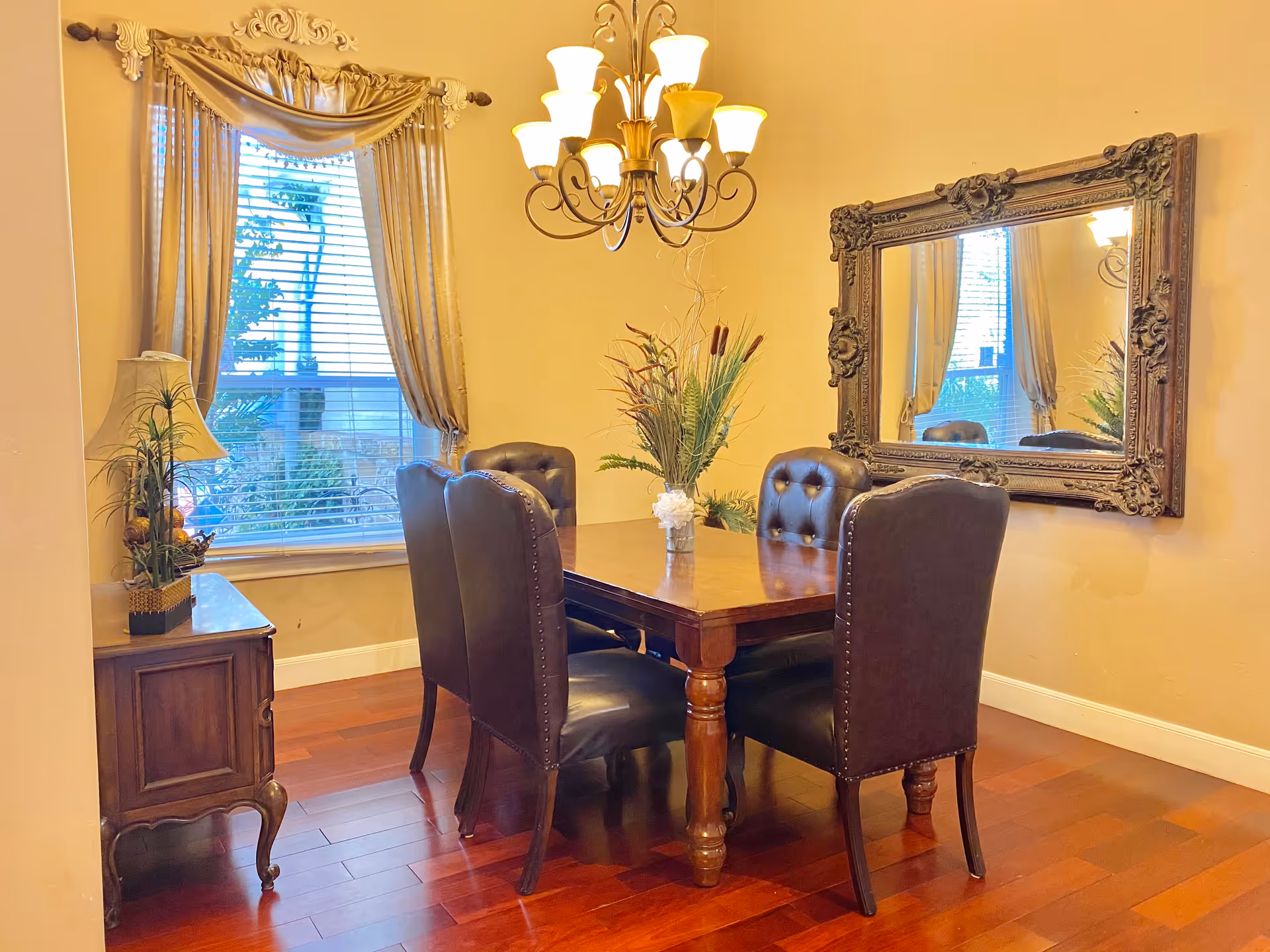 A dining room with a wooden table surrounded by six dark leather chairs. A decorative vase with plants is placed on the table. There is a large ornate mirror on the wall reflecting part of the room. A window with blinds and gold curtains is on the left side, next to a small wooden side table with a lamp and a plant. A chandelier with multiple lights hangs above the table. The floor is made of polished wood.