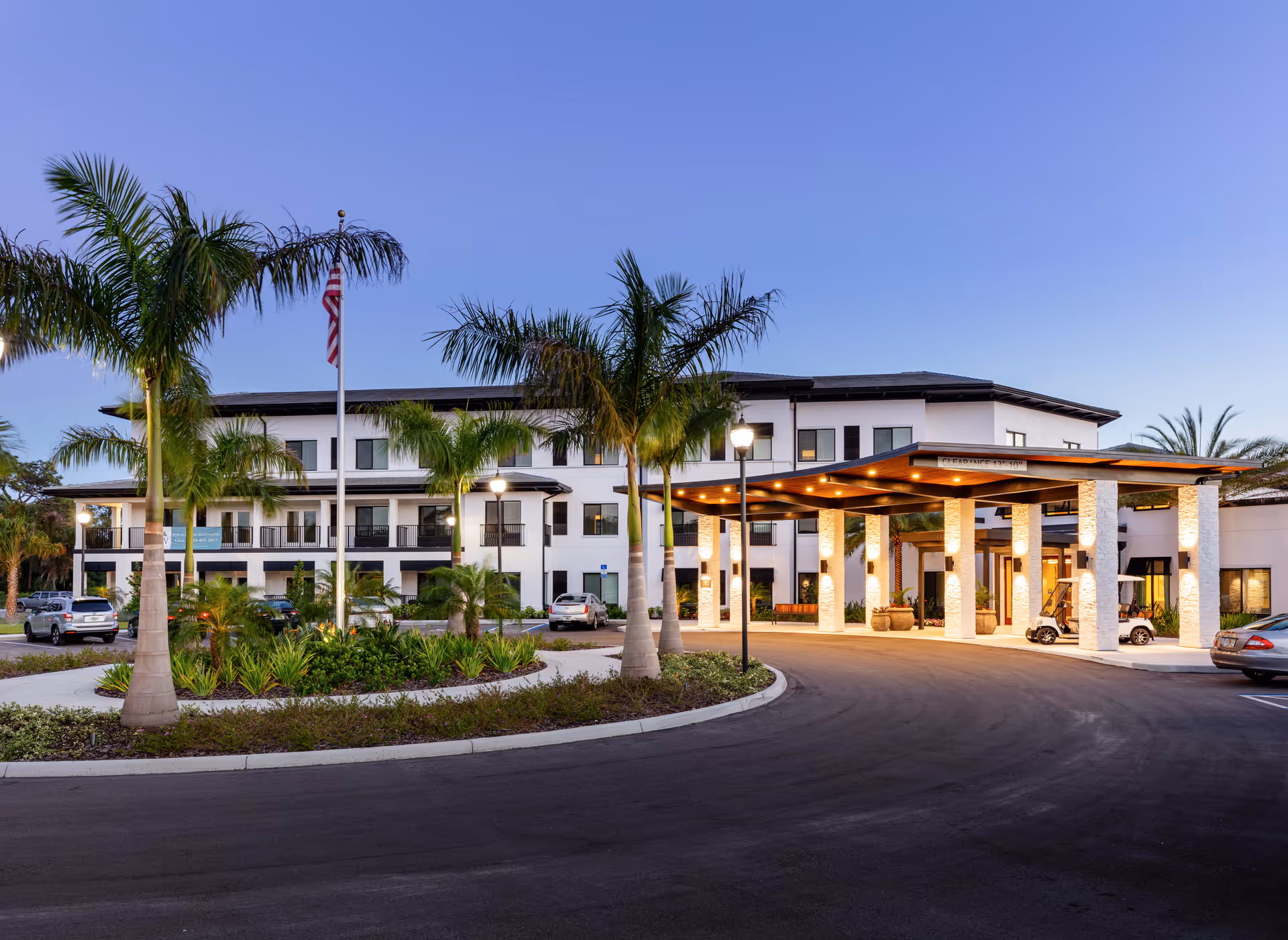 Exterior view of Amira Choice Sarasota senior living facility at dusk, showing a modern three-story building with a covered entrance, palm trees, landscaped roundabout, parked cars, and warm lighting.