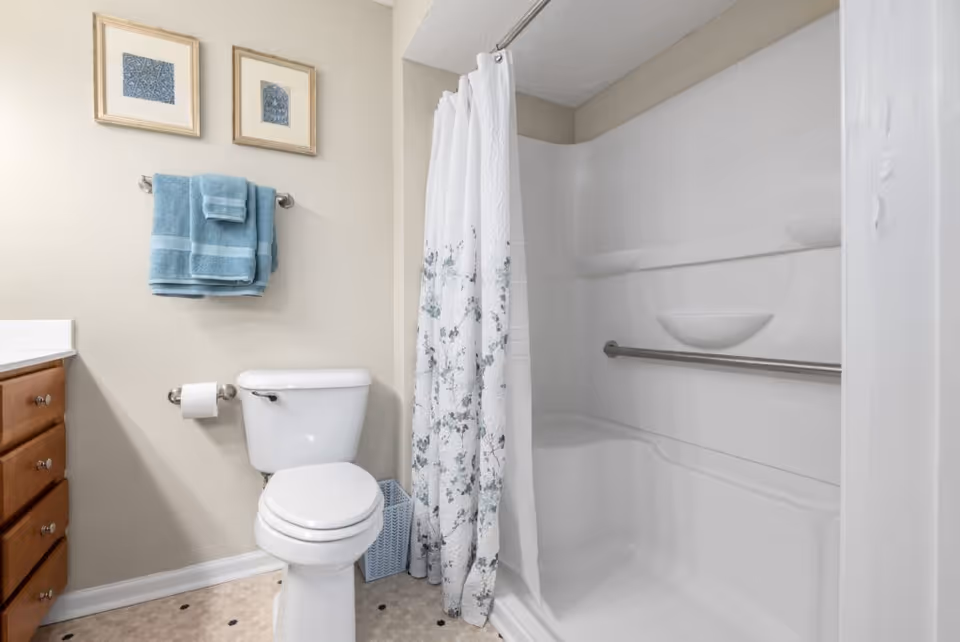 A bathroom with a white toilet, a shower with a white and floral shower curtain, a grab bar inside the shower, a wooden cabinet with drawers, two framed pictures on the wall, and blue towels hanging on a towel rack.