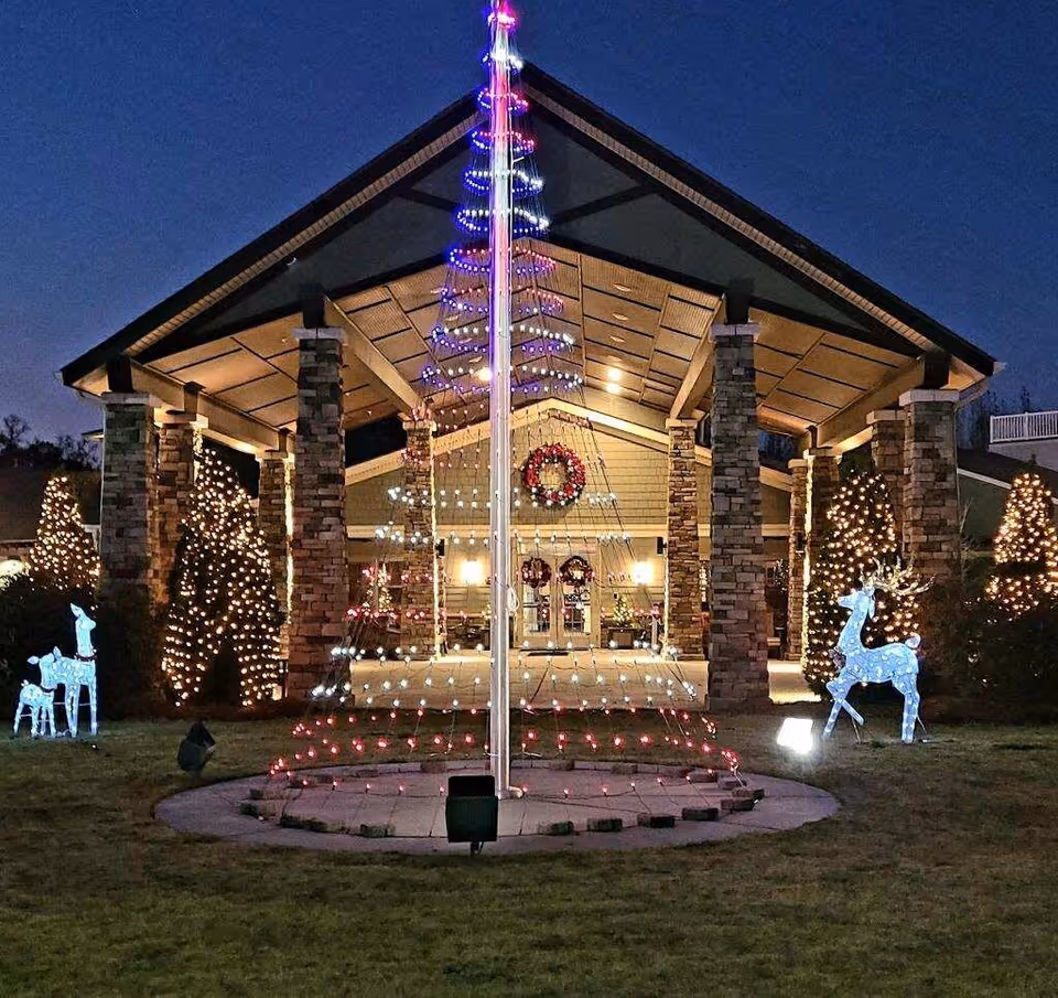 Entrance of a building at night decorated with Christmas lights, including a lighted tree structure around a flagpole, illuminated reindeer figures, and wreaths on the doors. The building has stone pillars and a covered porch area.