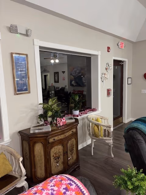 Interior view of a living room area in an assisted living facility featuring a wooden cabinet with decorative plants and items on top, two white chairs with yellow cushions, a large window opening into another room, and a door with an exit sign above it. The floor is wooden, and there are various decorations on the walls.