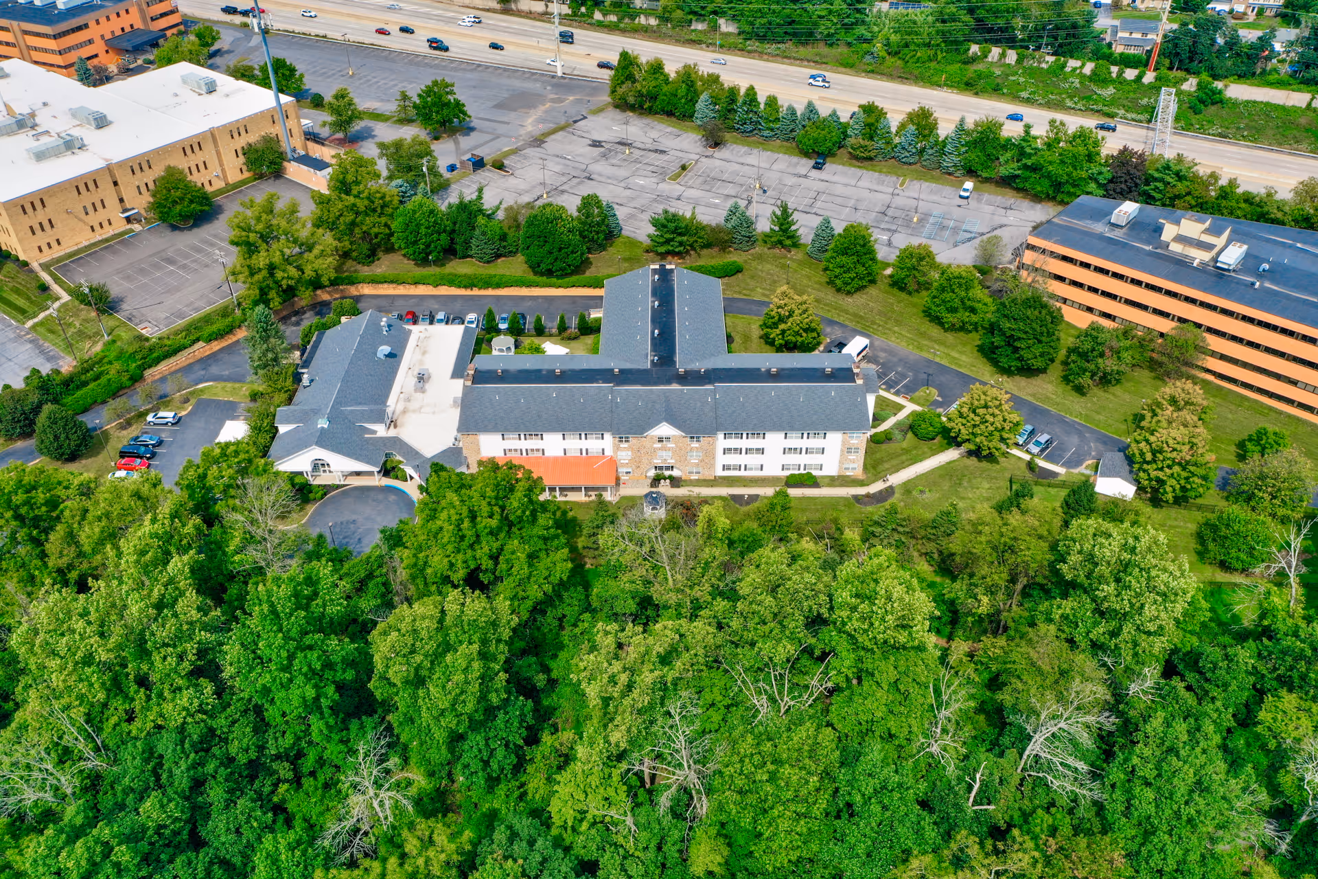 Aerial view of a senior living facility building surrounded by trees, lawns, parking lots, and nearby roads.