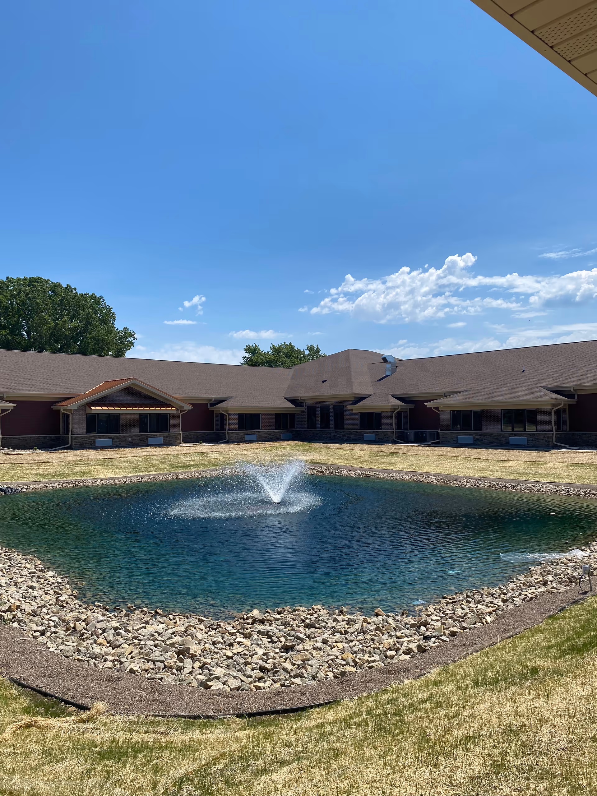 A courtyard area of CopperStone Assisted Living featuring a small pond with a central water fountain surrounded by rocks and grass, with a single-story building in the background under a clear blue sky.