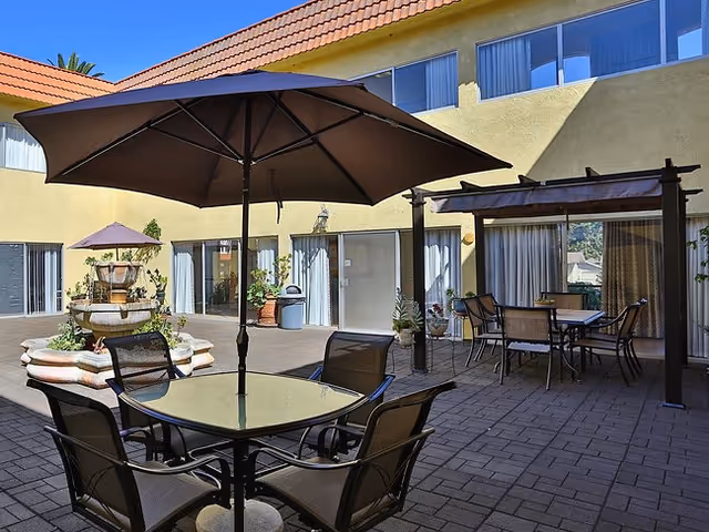 Sunlit courtyard with patio tables and umbrellas, a central fountain, and a pergola seating area.
