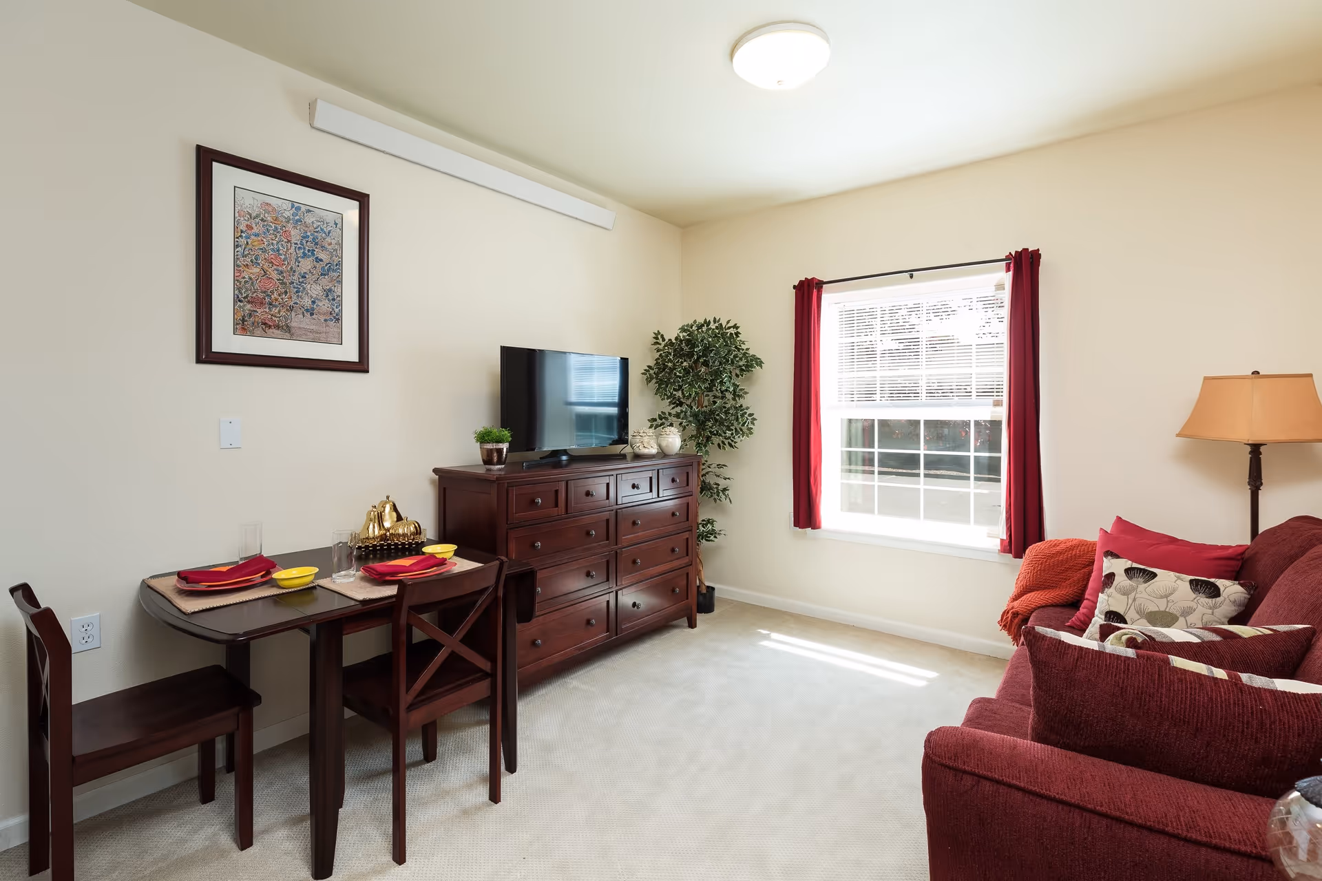 A cozy living room area with a maroon sofa adorned with patterned and solid pillows, a floor lamp, a window with red curtains, a wooden dresser with a flat-screen TV and decorative items, a small dining table set for two with red napkins and yellow bowls, and a framed floral artwork on the wall.
