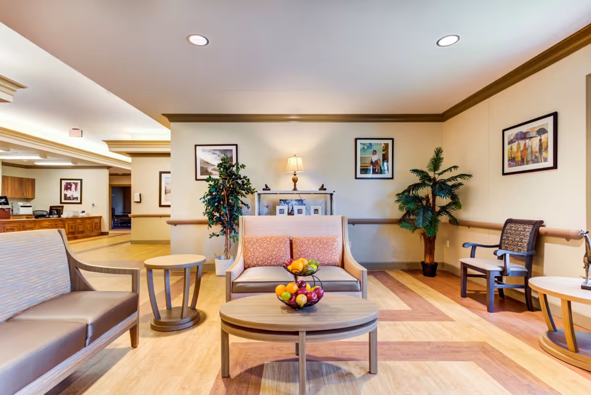 Well-lit senior living common room with seating, coffee tables, artwork, and potted plants.