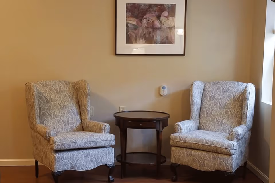Two patterned armchairs with wooden legs positioned on either side of a round wooden side table against a beige wall. Above the table hangs a framed floral artwork. A window is visible on the right side, allowing natural light into the room.