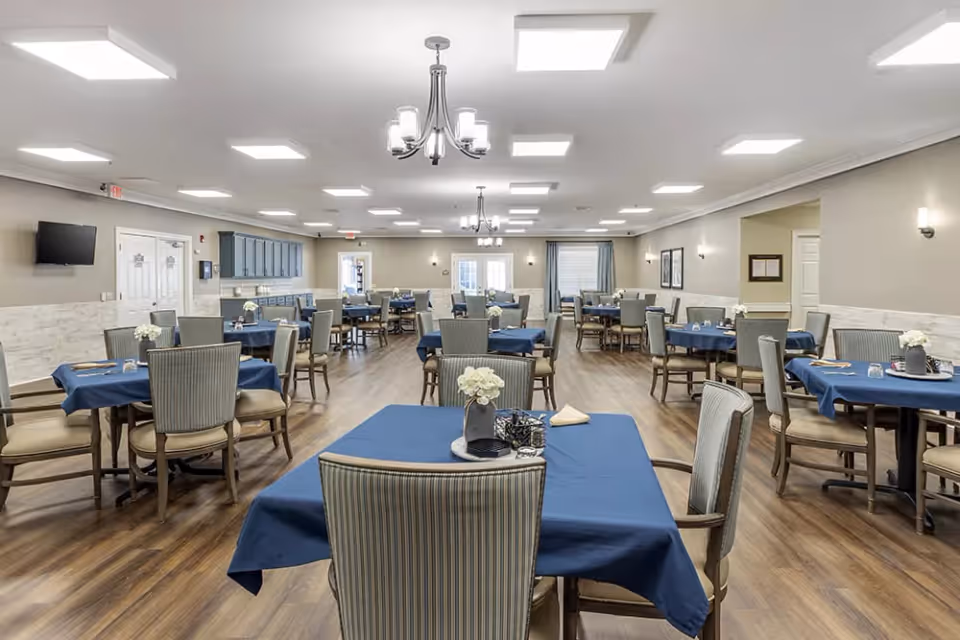 A spacious dining room with multiple tables covered in blue tablecloths, each set with chairs and small floral centerpieces. The room has wood flooring, neutral-colored walls, ceiling lights, and chandeliers. There is a wall-mounted TV and cabinetry along one side of the room.