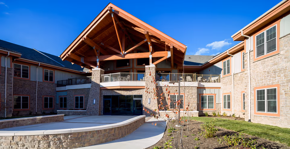 Front exterior of a senior living facility with a stone facade, large timber-covered entrance, windows, and landscaped walkway.