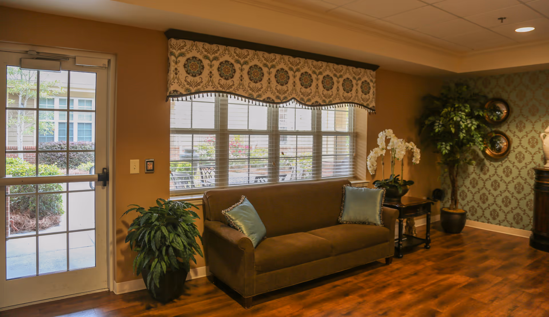 Comfortable seating area with a brown sofa, decorative pillows and plants beside a window and glass door in a senior living facility.