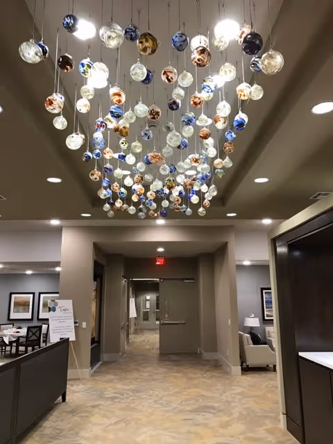 Interior view of a senior living facility hallway with a decorative ceiling installation featuring numerous hanging glass orbs in various colors. The hallway leads to a door at the far end, with seating areas and framed artwork visible on the walls to the sides.