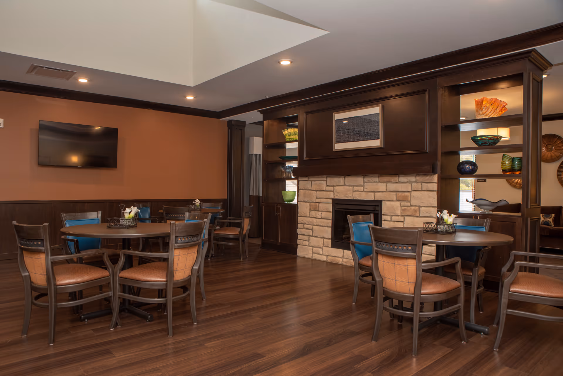 A cozy interior common area with round tables and chairs arranged on a wooden floor. The room features a stone fireplace with dark wood cabinetry and shelves displaying decorative glass bowls. A flat-screen TV is mounted on the brown wall, and recessed ceiling lights illuminate the space.