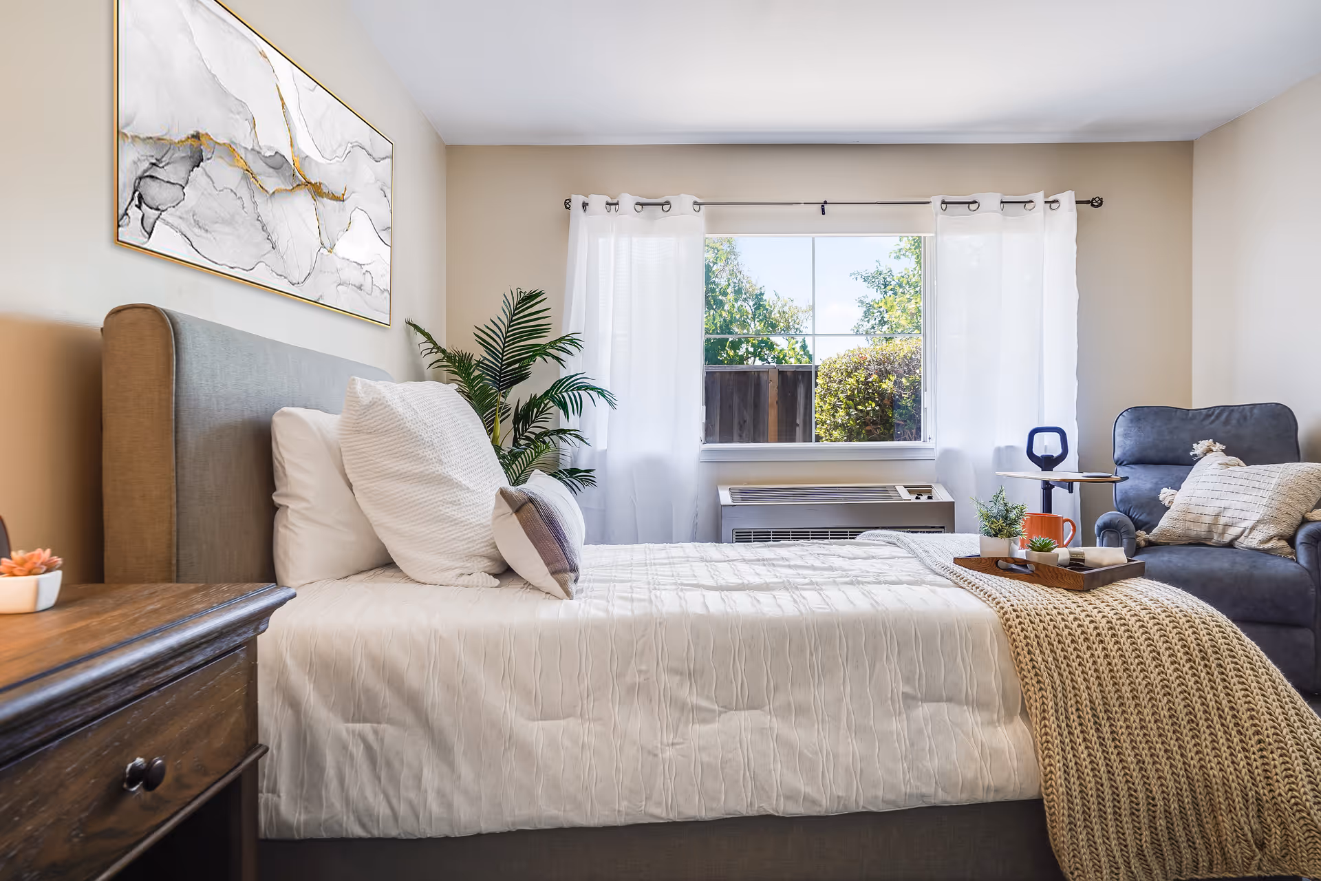 A bright, neatly made bedroom featuring a bed with pillows and a knit throw, a wooden nightstand, an armchair, a potted plant, wall art, and a window with white curtains.