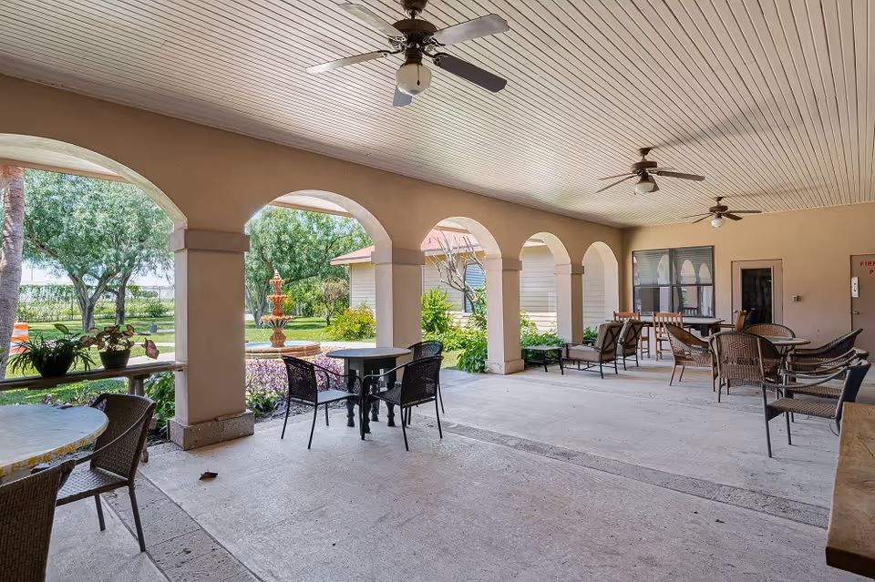 Covered outdoor patio with arched openings, multiple tables and chairs, ceiling fans, and a view of a garden fountain.