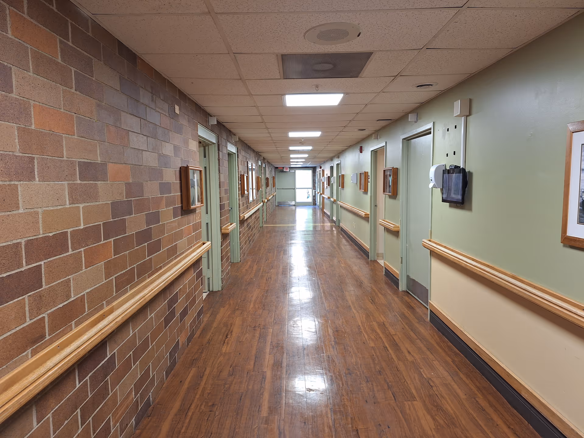 A long, empty hallway in a senior living facility with wood-patterned flooring, brick wall on the left, and light green painted walls on the right. Several doors line both sides of the hallway, with handrails running along the walls. The ceiling has recessed lighting panels and a drop ceiling design.