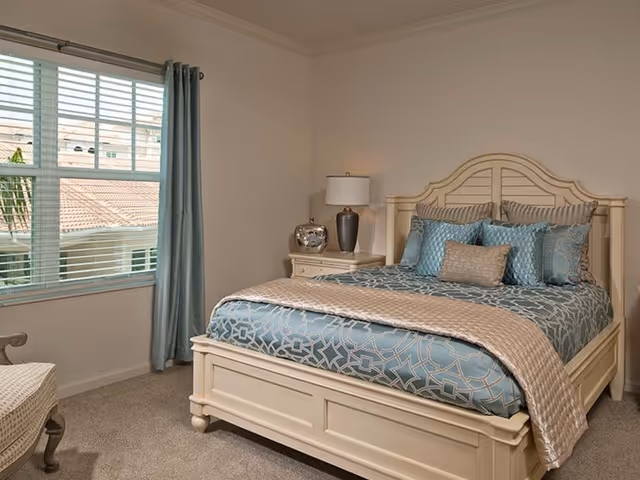 A neatly made bed with a cream-colored wooden frame and headboard, adorned with blue and beige patterned bedding and multiple pillows. Next to the bed is a cream-colored nightstand with a silver decorative item and a lamp with a white shade. A window with white blinds and blue curtains is on the left side, allowing natural light into the room. The floor is carpeted in a neutral tone.