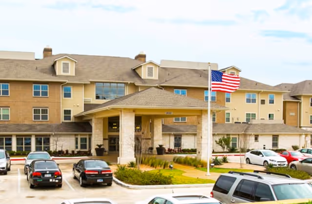 Exterior view of a multi-story retirement resort building with a covered entrance, several parked cars in the parking lot, an American flag on a flagpole, and landscaped greenery around the entrance.