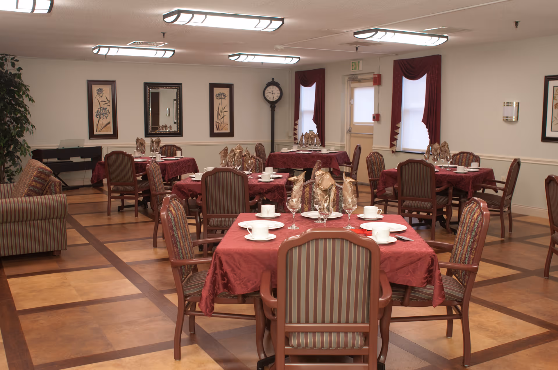 A dining room with multiple tables covered in red tablecloths, each set with white plates, cups, and folded napkins. The room has wooden chairs with striped upholstery, framed floral artwork on the walls, a tall clock near the windows with red curtains, and a striped couch on the left side.