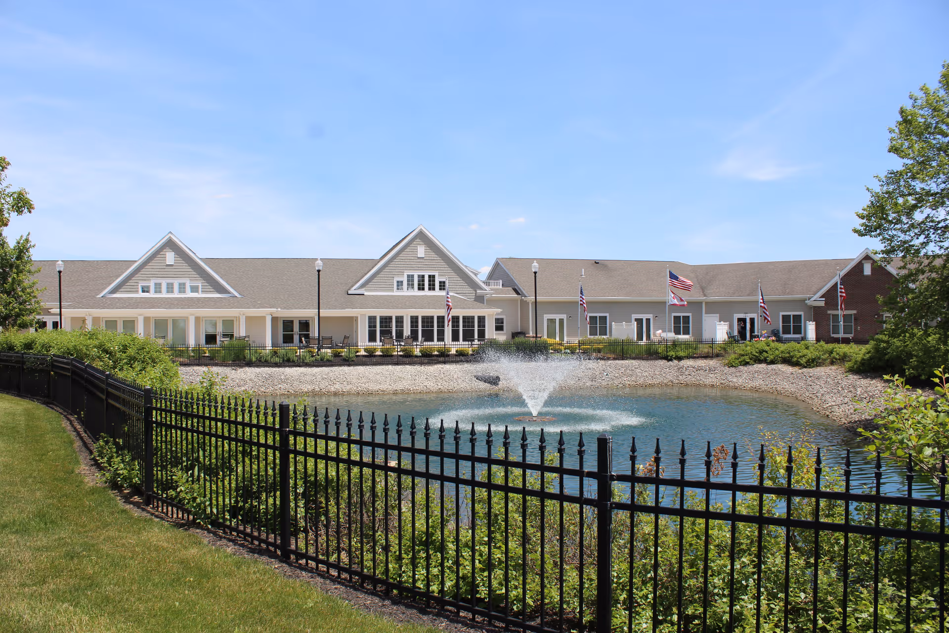 View of a senior living facility building named Briarwood Village with a pond and a water fountain in front, surrounded by a black metal fence and greenery under a clear blue sky.