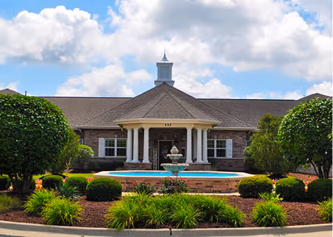 Front exterior view of a single-story brick building with a peaked roof and a small cupola on top. The entrance features white columns and a covered porch. In front of the building is a circular fountain surrounded by neatly trimmed bushes and landscaped greenery under a partly cloudy sky.