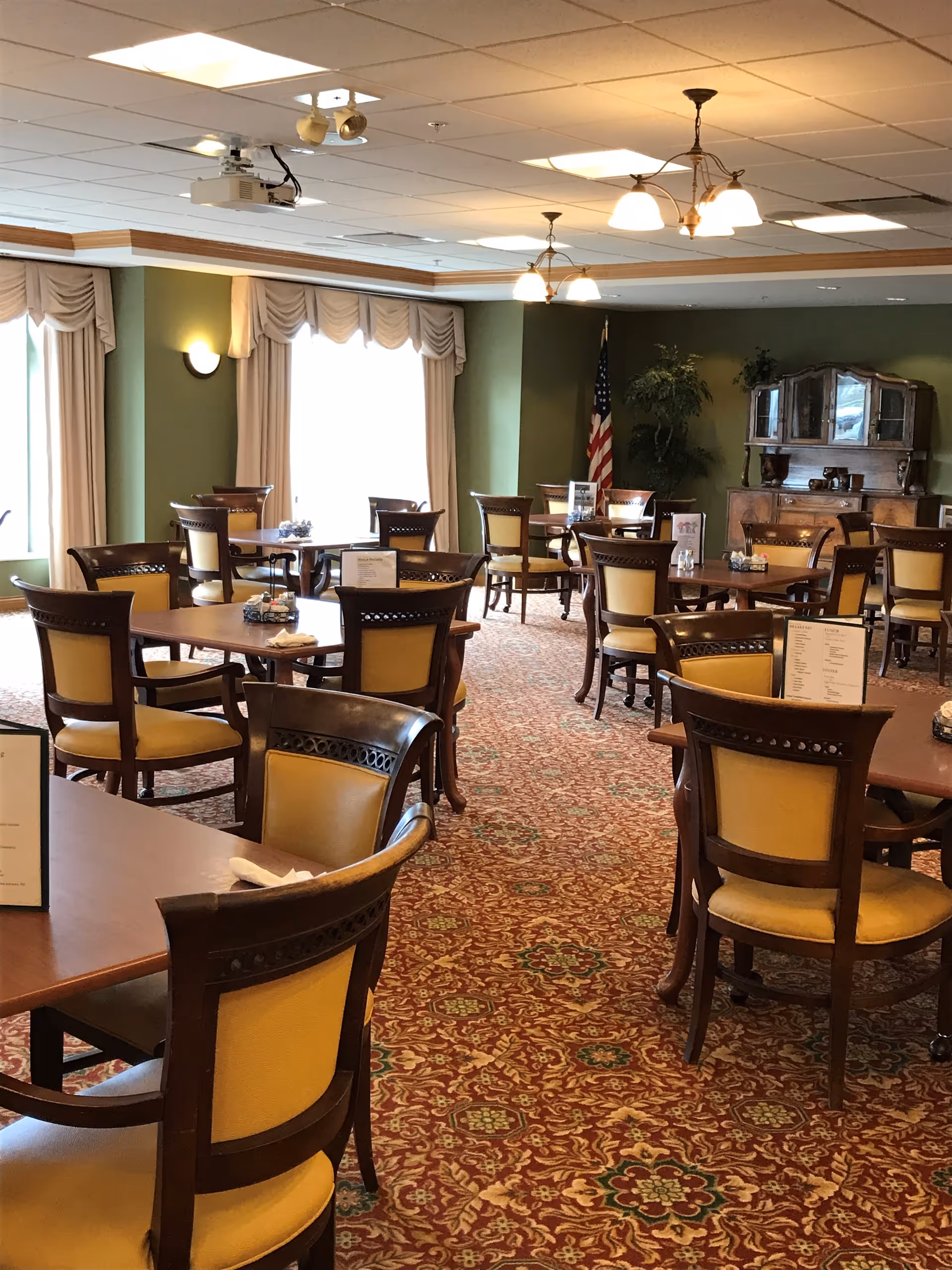 A dining room with multiple wooden tables and chairs with yellow cushions. The room has green walls, large windows with beige curtains, a patterned carpet, and ceiling lights. There is an American flag and a wooden cabinet with decorative items in the background.