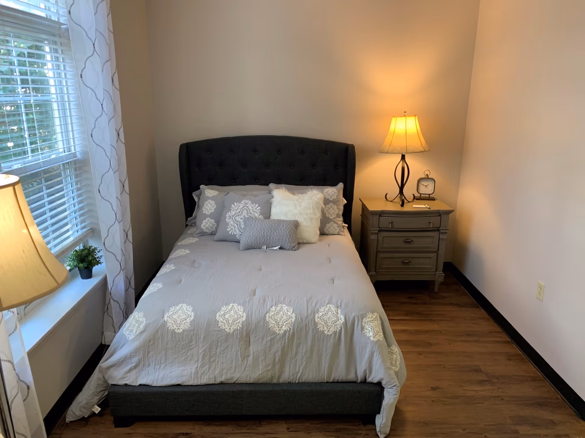 A neatly made bed with patterned bedding and pillows between a window and a nightstand with a lamp in a small bedroom.