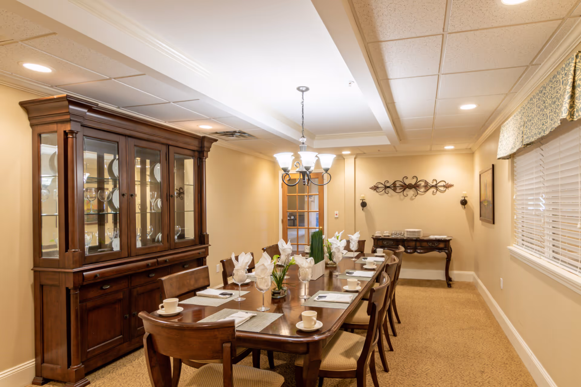 A formal dining room with a long wooden table set for eight people, including white napkins, cups, and glasses. There is a wooden china cabinet on the left filled with plates and glassware, a chandelier hanging above the table, and a window with blinds and a valance on the right. The walls are beige with decorative wall art and a side table at the far end of the room.