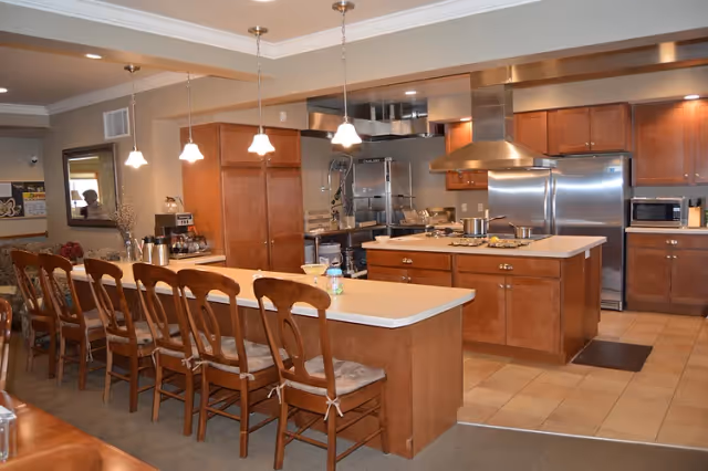 A spacious kitchen area with a long island counter featuring six wooden chairs with cushions. The kitchen has wooden cabinets, stainless steel appliances including a refrigerator and stove with a range hood, and pendant lights hanging above the island. The floor is tiled and the overall design is warm and inviting.