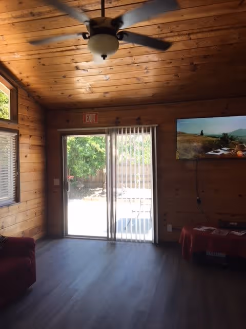 Interior view of a wooden-paneled room with a ceiling fan, a sliding glass door with vertical blinds leading outside, a wall-mounted flat screen TV showing a landscape scene, a red armchair on the left, and a small table covered with a red cloth on the right.