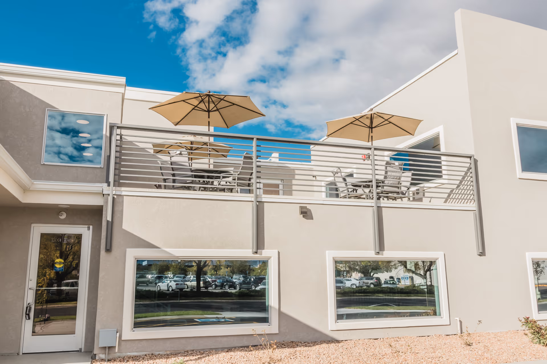 Exterior view of a modern building with a second-floor balcony patio furnished with tables and umbrellas.