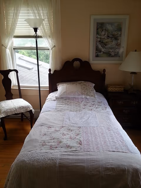 A small bedroom with a single bed covered in a patchwork quilt with floral patterns. Next to the bed is a wooden nightstand with a lamp and a small box. A framed painting hangs on the wall above the nightstand. To the left of the bed is a wooden chair with a white cushion, and a tall floor lamp stands in front of a window with sheer white curtains.