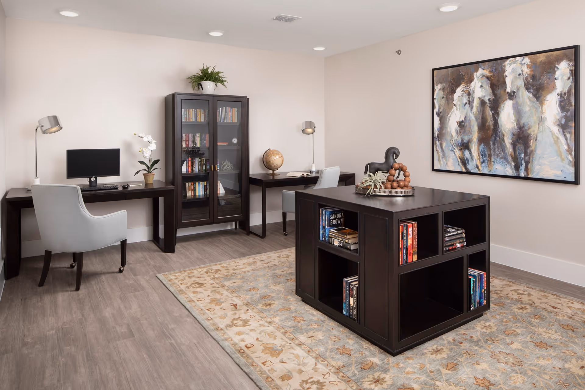 A quiet, well-lit room with two desks each equipped with a lamp and chair, a computer monitor on one desk, a glass-front bookcase filled with books, and a central dark wood cube bookshelf with books and decorative items. A large painting of white horses hangs on the wall, and a patterned area rug covers part of the wooden floor.
