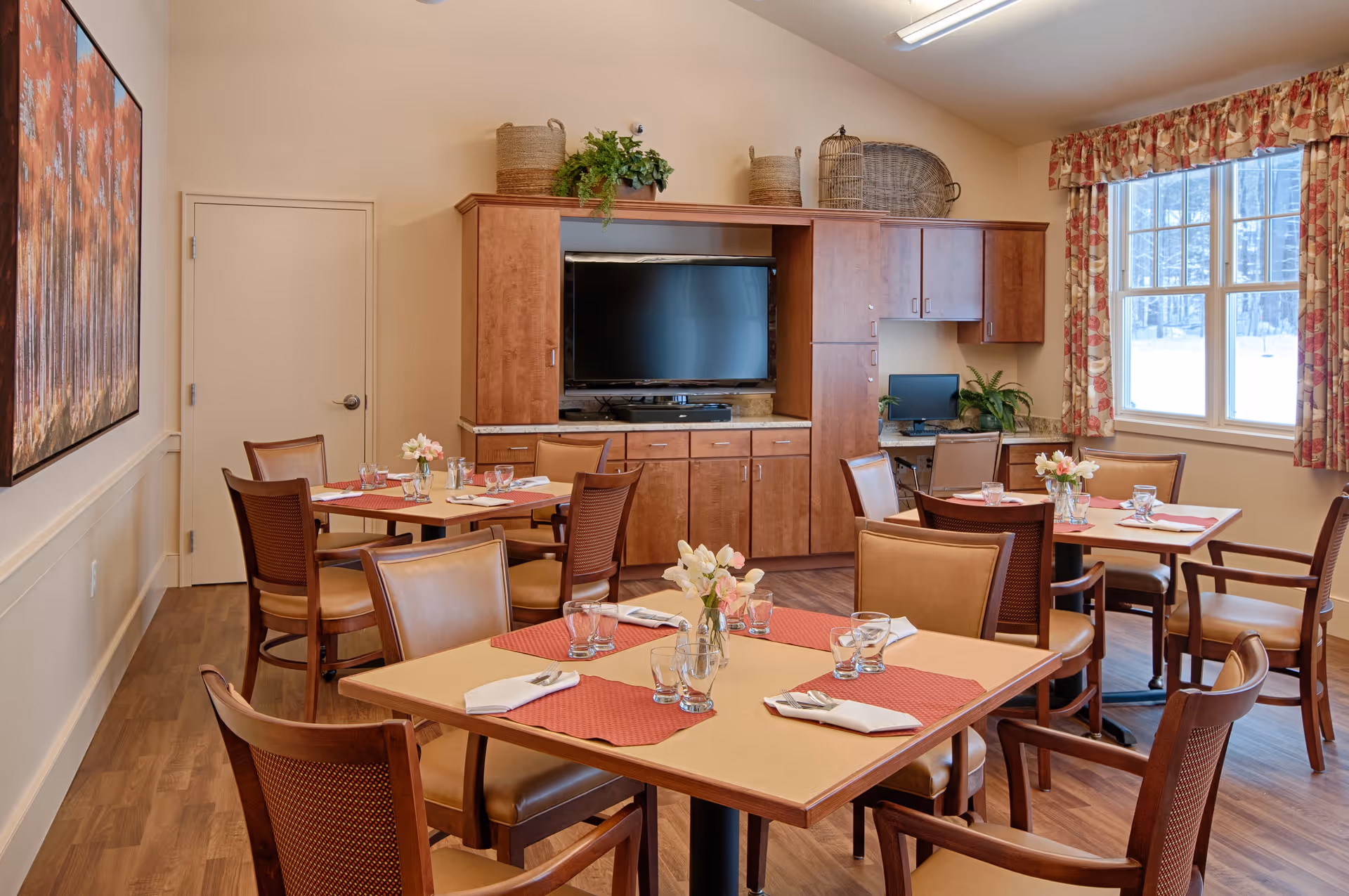 A cozy dining room in a senior living facility with several tables set with placemats, napkins, glasses, and small flower vases. The room features wooden chairs with cushioned seats, a large flat-screen TV mounted on a wooden cabinet, a computer workstation in the corner, and a window with floral curtains letting in natural light.