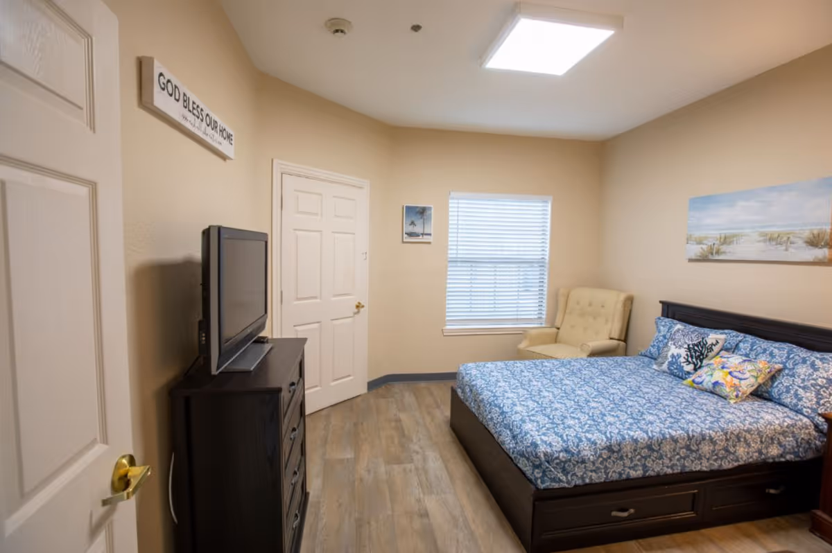 A cozy bedroom with a bed covered in blue and white patterned bedding, a beige armchair in the corner, a dark wooden dresser with a flat-screen TV on top, and a window with blinds. The walls are beige with a beach-themed painting and a sign that reads 'GOD BLESS OUR HOME'.