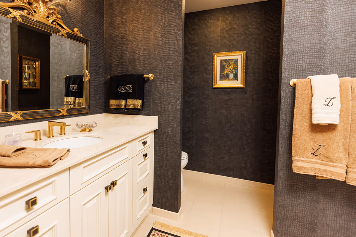 A stylish bathroom with a white vanity and gold fixtures against dark textured walls, a framed mirror, tan monogrammed towels, and a tucked-away toilet.